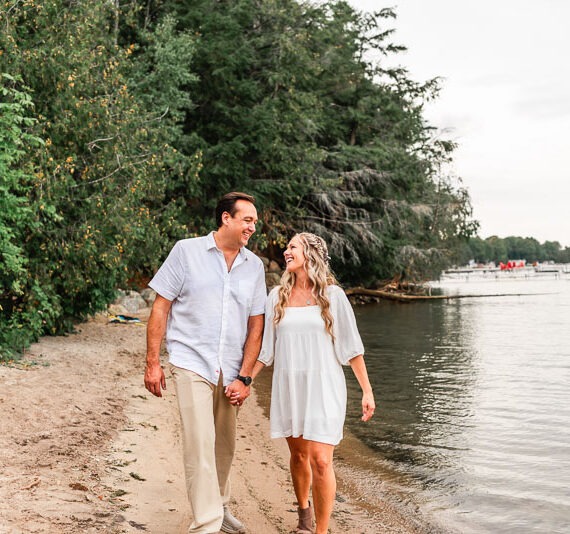 Engaged couple walking along the beach of a lake.