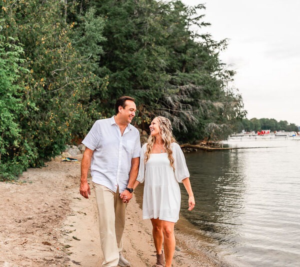 Engaged couple walking along the beach of a lake.